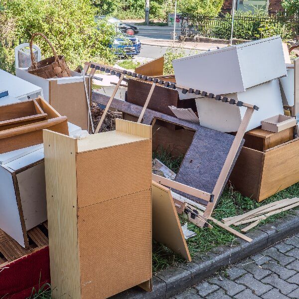 A chaotic pile of broken and discarded furniture on a sidewalk curb, including cabinets, drawers, and shelves.